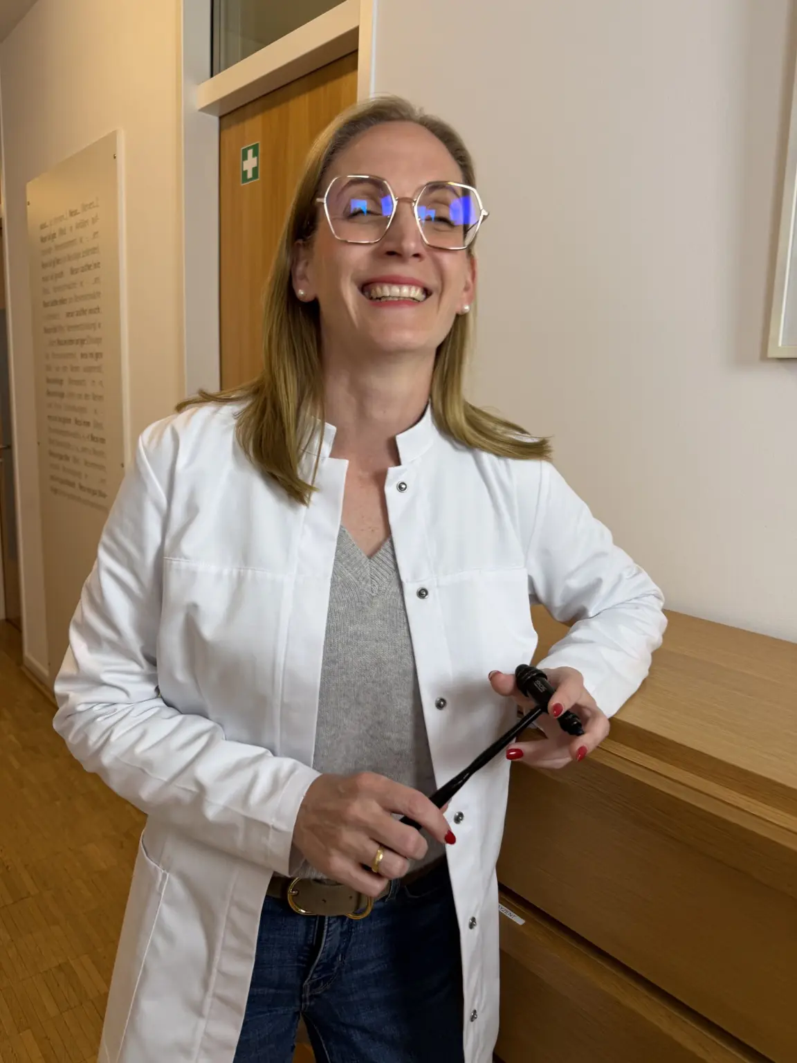 Prof. Dr. med. Anke Lührs lacht, im weißen Kittel mit Reflexhammer in der Hand, im Flur der Praxis